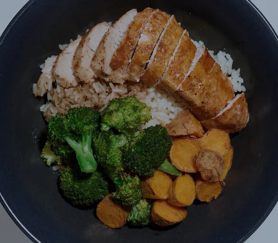 An athlete’s meal of sliced chicken, rice, broccoli and sweet potato in a dark bowl